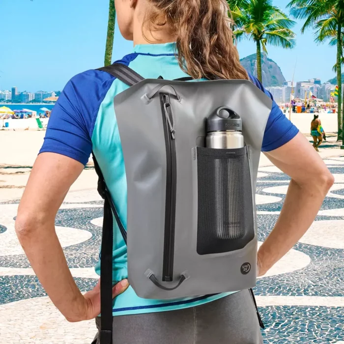 Frau spaziert entspannt am Strandboulevard entlang – auf dem Rücken ein moderner, grauer wasserdichter Rucksack mit Netztasche, aus der eine Edelstahl-Trinkflasche hervorschaut.