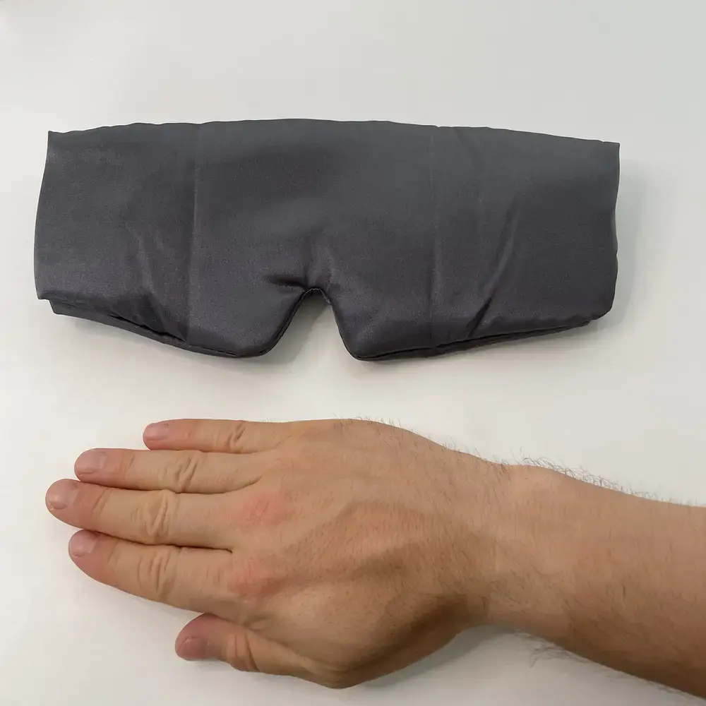 Kitsch Schlafmaske aus grauem Satin flach auf Tisch mit Hand zum Grössenvergleich