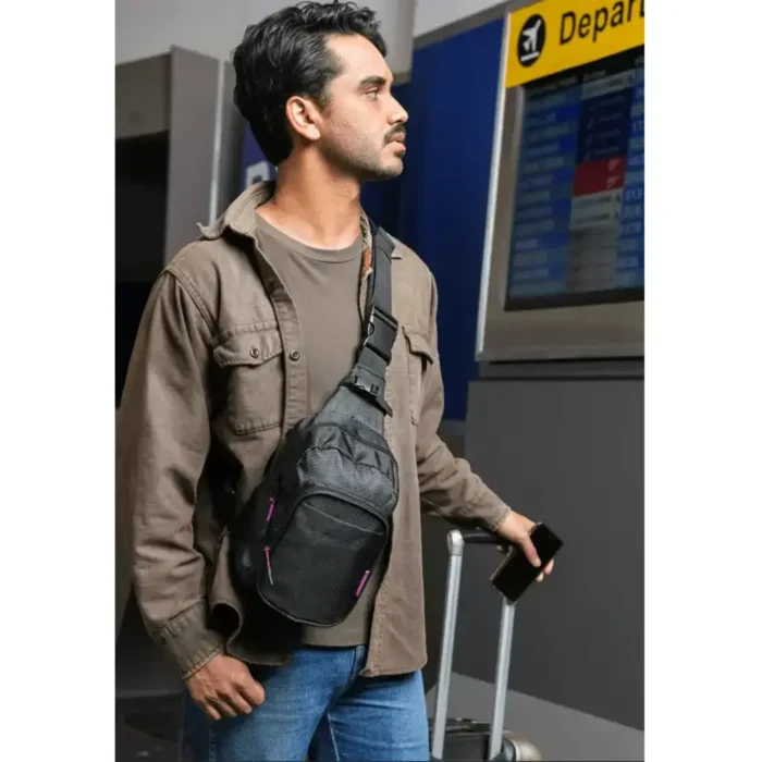 Mann am Flughafen mit schwarzem Sling Pack und Rollkoffer, blickt auf die Abflugtafel.