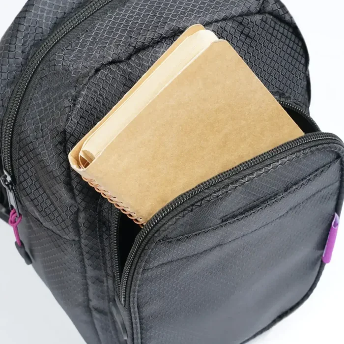 Nahaufnahme eines schwarzen Sling Packs mit geöffnetem Frontfach, aus dem ein braunes Notizbuch herausragt.