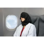 Frau sitzt im Flugzeug am Fenster und trägt eine schwarze Schlafkapuze über Kopf und Augen, um ungestört zu schlafen. Der Blick geht auf den Himmel durch das Flugzeugfenster.