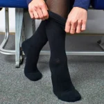 Person im Flugzeugsitz zieht sich schwarze Reisesocken über eine Strumpfhose, die Beine stehen auf einem Teppichboden im Kabinenbereich.