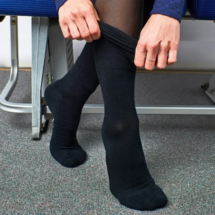 Person im Flugzeugsitz zieht sich schwarze Reisesocken über eine Strumpfhose, die Beine stehen auf einem Teppichboden im Kabinenbereich.