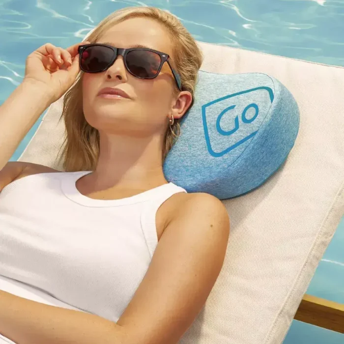 Frau mit Sonnenbrille liegt entspannt auf einer Liege am Pool, den Kopf auf einem blauen Strandkissen der Marke Go Travel.