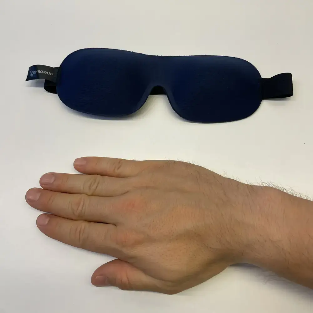 OHROPAX Schlafmaske neben einer Hand zur Grössenveranschaulichung auf weissem Untergrund